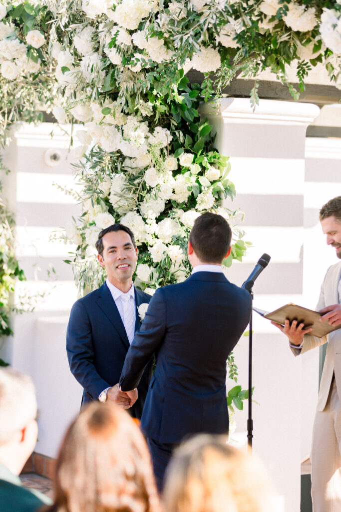 Two grooms getting married in front of triple wedding arch at Hotel Californian Sirena Rooftop with white flowers. Gay wedding in Santa Barbara.