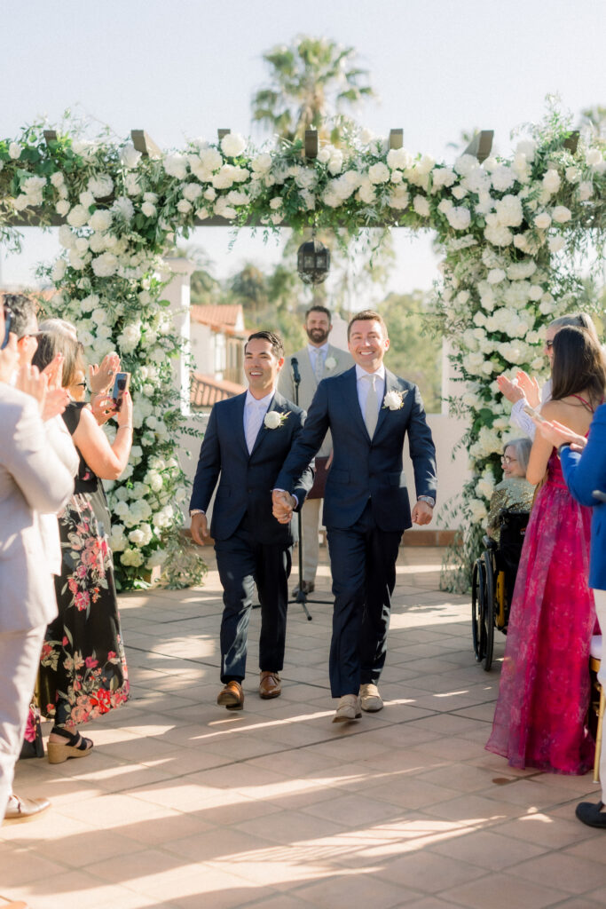 Two grooms getting married in front of triple wedding arch at Hotel Californian Sirena Rooftop with white flowers. Gay wedding in Santa Barbara.