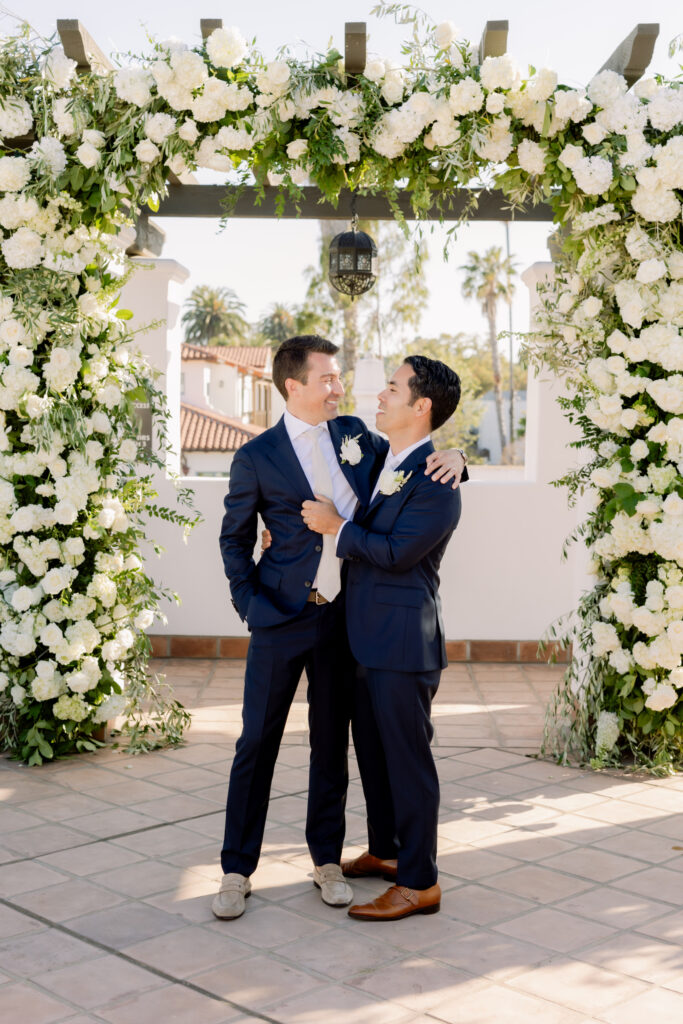 Two grooms getting married in front of triple wedding arch at Hotel Californian Sirena Rooftop with white flowers. Gay wedding in Santa Barbara.