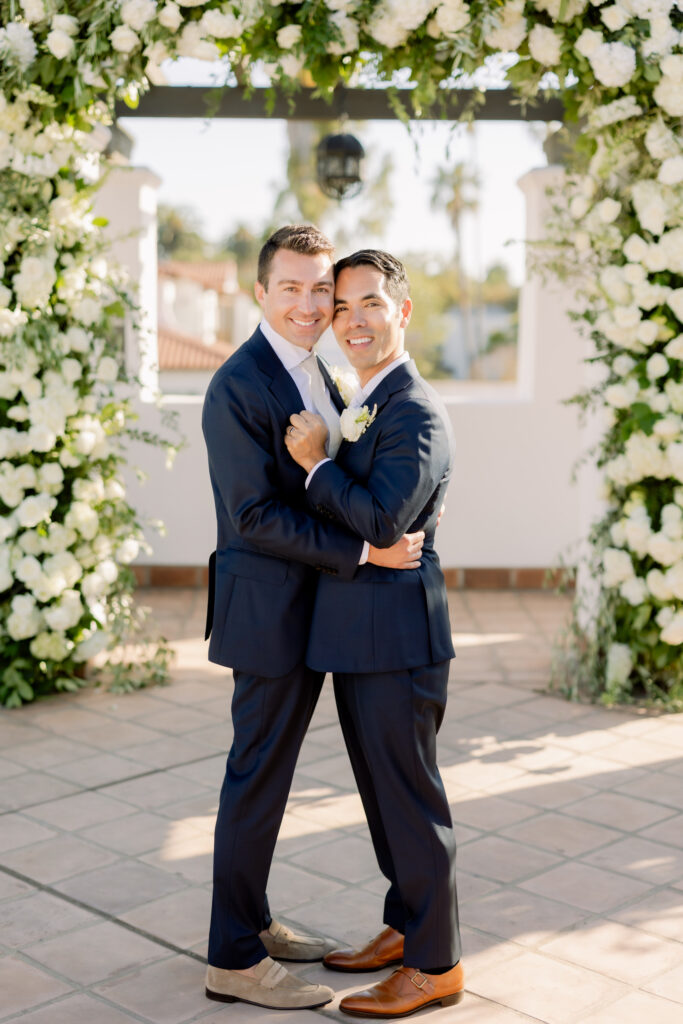 Two grooms getting married in front of triple wedding arch at Hotel Californian Sirena Rooftop with white flowers. Gay wedding in Santa Barbara.