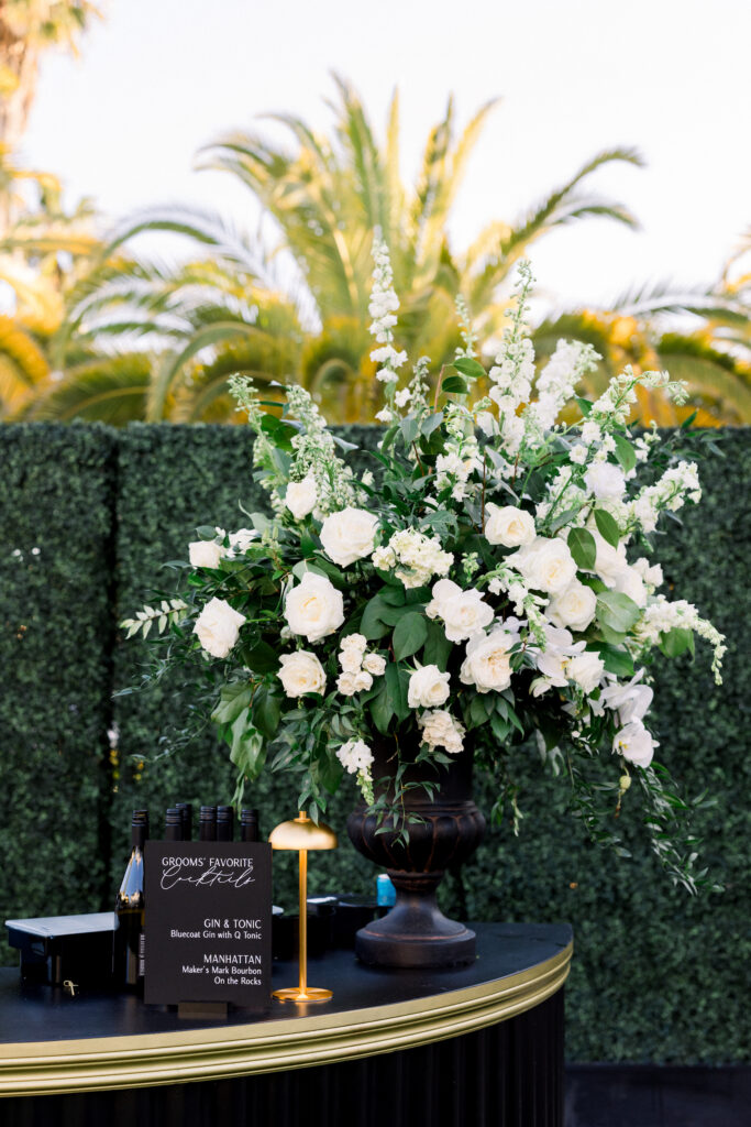 Bar urn at cocktail hour with white flowers, table lamp and hedge wall