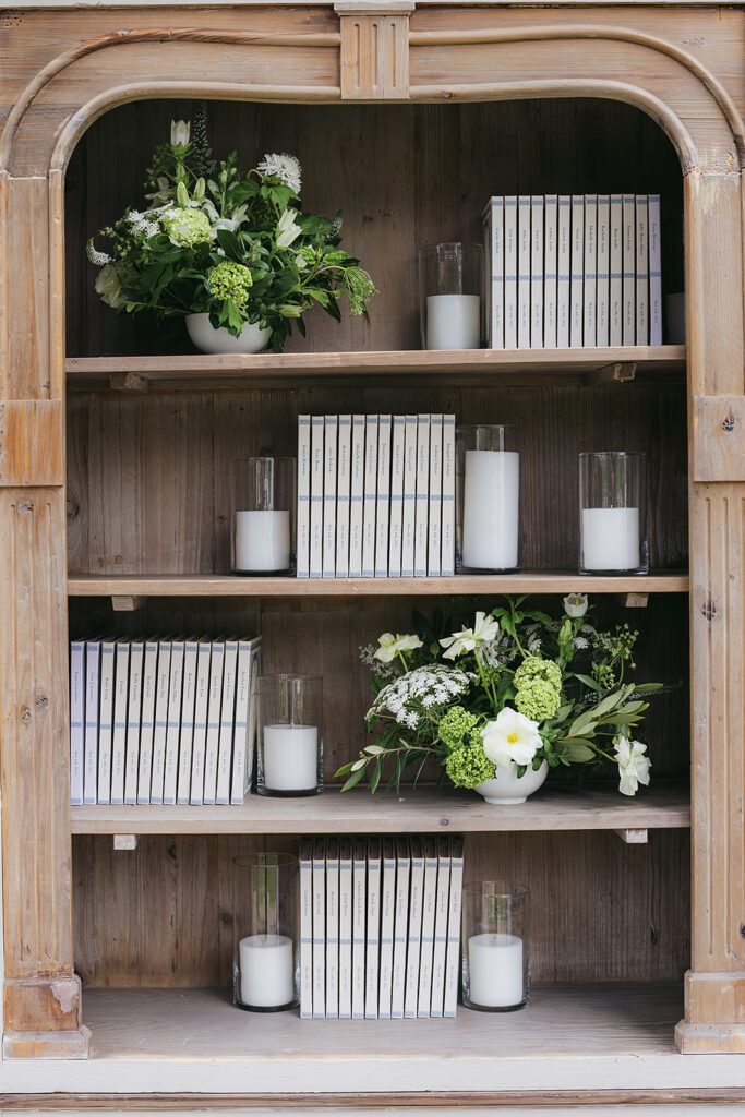 Wedding Escort card display using a bookshelf and custom journals for guests with candles and small floral arrangements on the bookshelves next to books