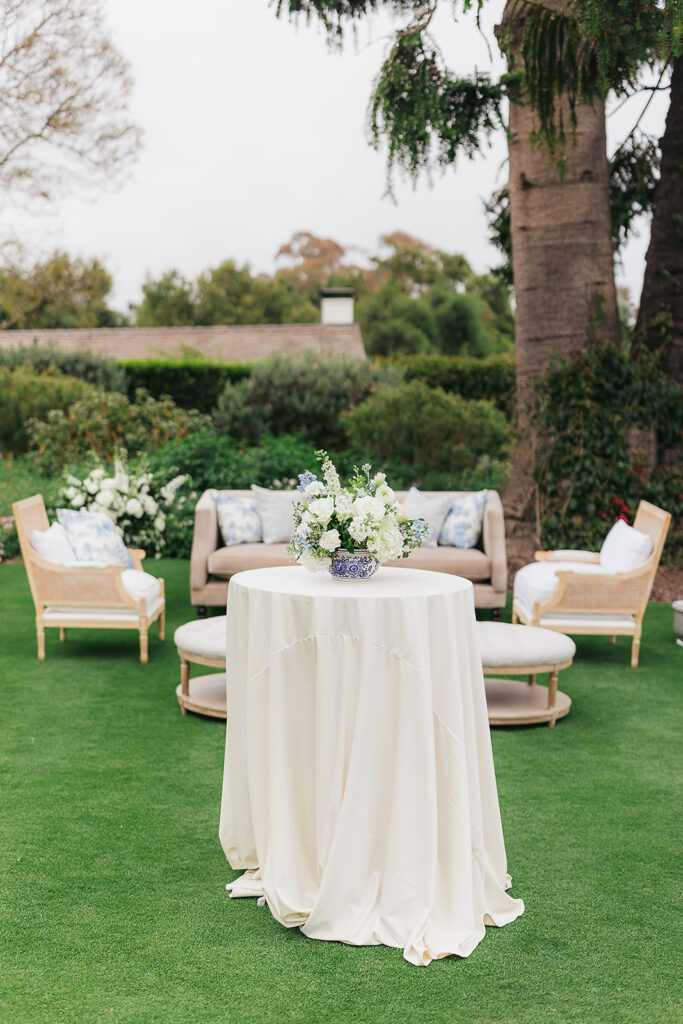 Tall cocktail table with floral centerpiece with white and blue flowers in blue and white ginger jars on neutral linens with lounge in background