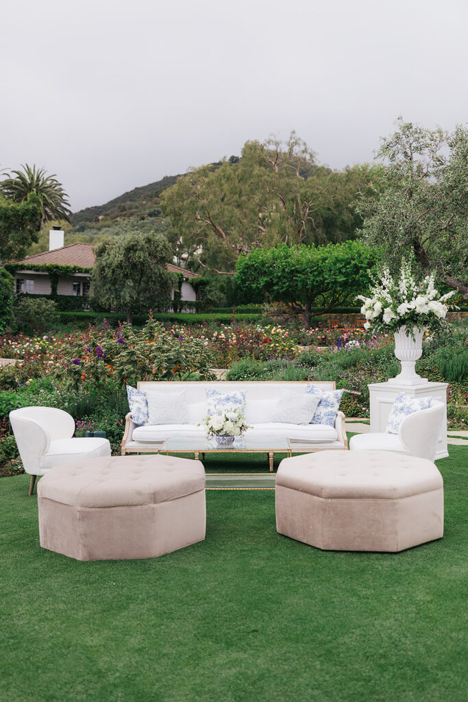 Neutral and elegant upholstered lounge with blue and white throw pillows in garden at wedding with large floral urn on white pillar