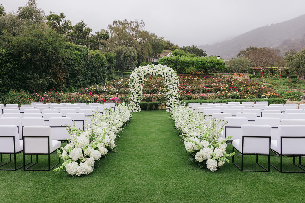 Wedding at San Ysidro Ranch in Montecito, California. Rounded wedding arch with lots of white flowers and a floral aisle. White upholstered ceremony chairs. 