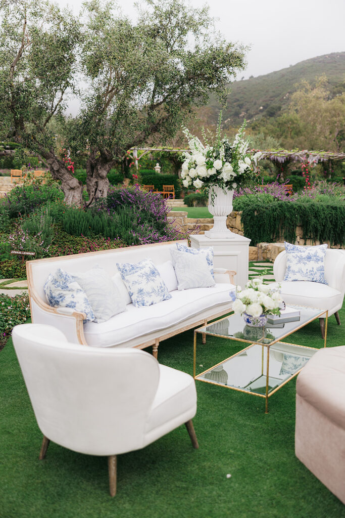 Neutral and elegant upholstered lounge with blue and white throw pillows in garden at wedding with large floral urn on white pillar