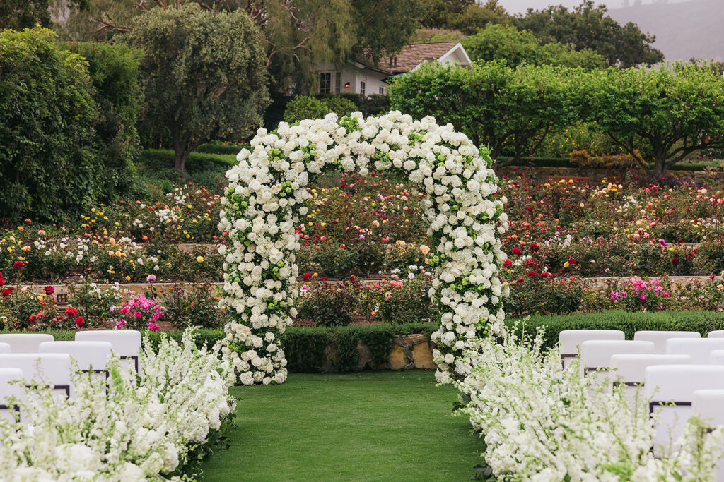 Wedding at San Ysidro Ranch in Montecito, California. Rounded wedding arch with lots of white flowers and a floral aisle. White upholstered ceremony chairs. Colorful rose garden in the background.