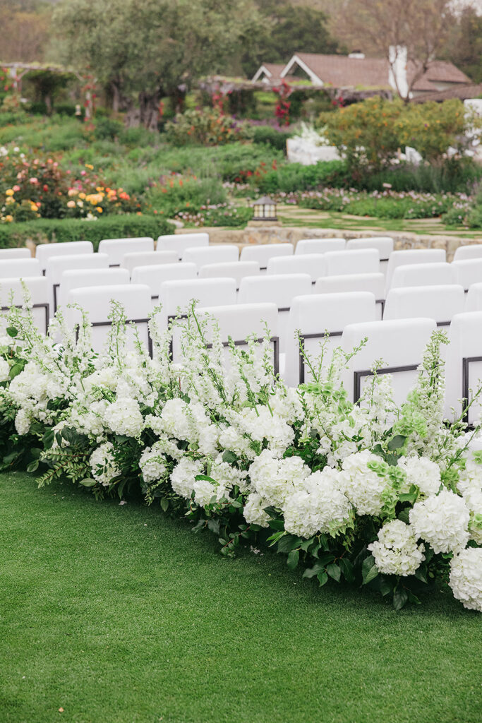 White upholstered wedding ceremony chairs with lush and whimsical white floral aisle at San Ysidro Ranch in Montecito, California.