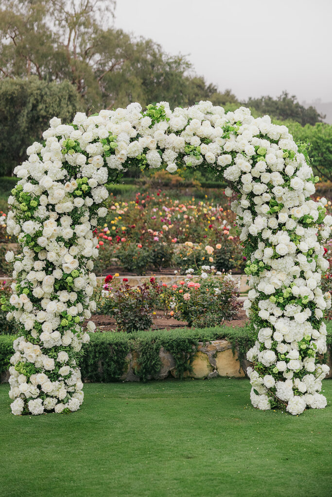 Ceremony at San Ysidro Ranch in Montecito, California. Rounded wedding arch with lots of white flowers. Colorful rose garden in the background.