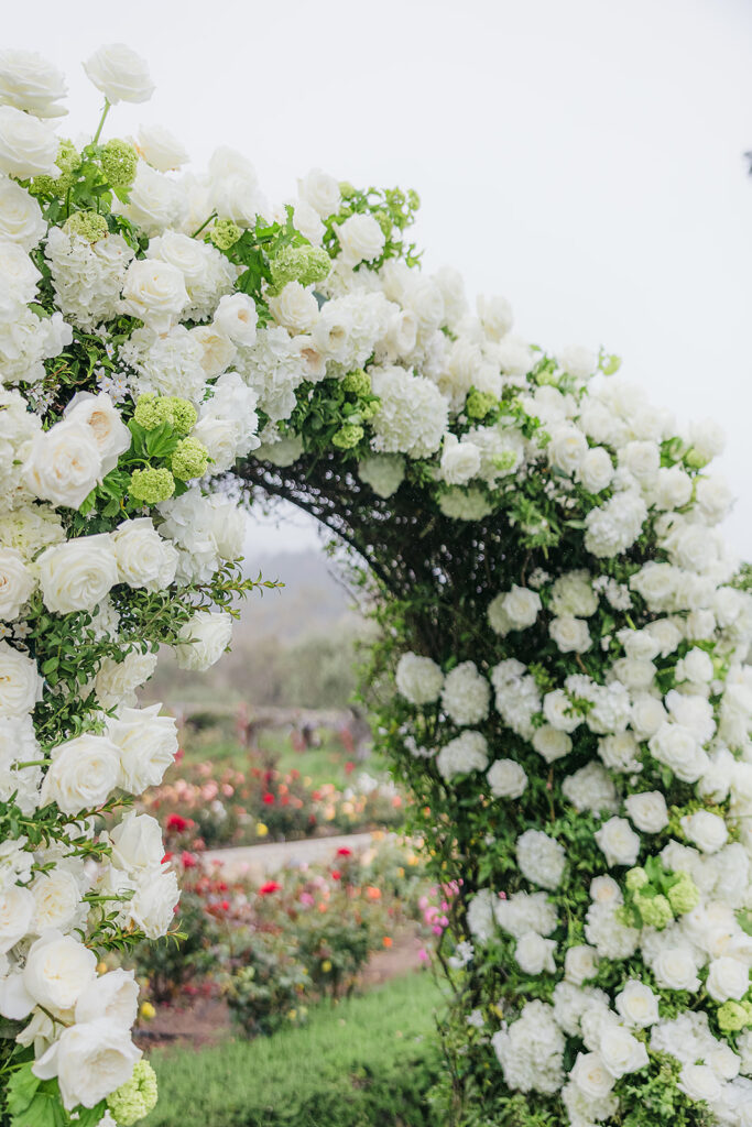 Ceremony at San Ysidro Ranch in Montecito, California. Rounded wedding arch with lots of white flowers. Colorful rose garden in the background.