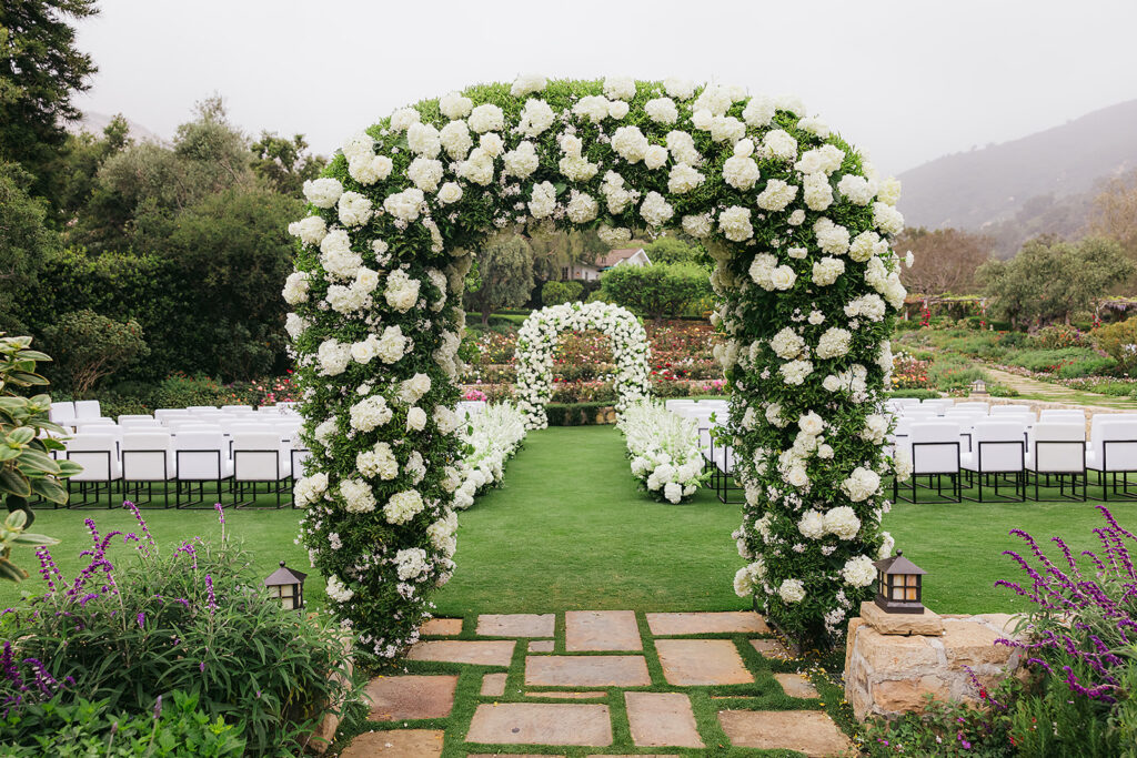 Double wedding arch with white flowers and a floral aisle. White upholstered ceremony chairs. Wedding at San Ysidro Ranch in Montecito, California.