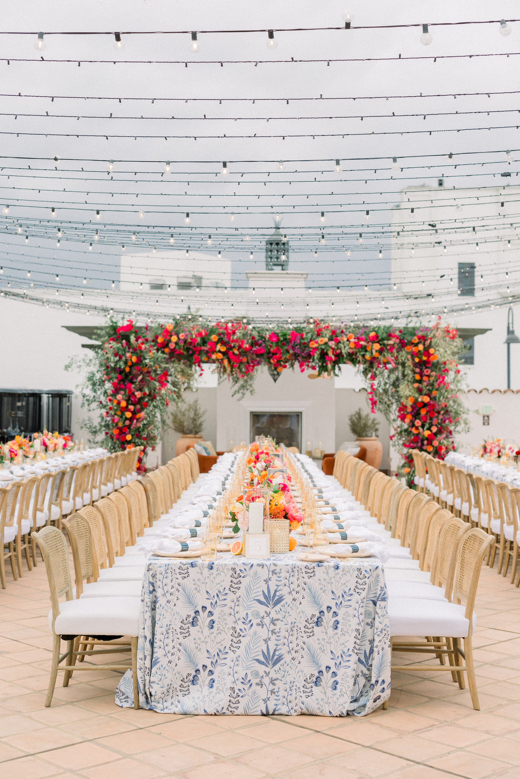 Welcome party dinner on rooftop with blue patterned linens, colorful flowers on long feasting tables, and large floral installation overhead with string lights