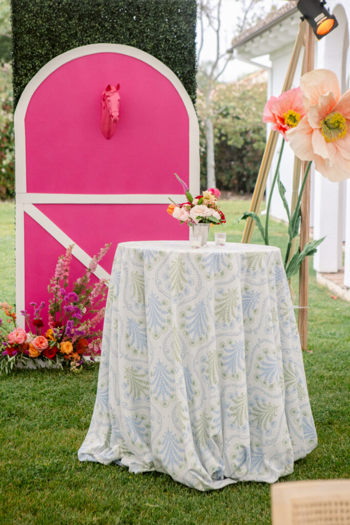 Cocktail table with green patterned linen and small colorful floral arrangement in mint julep cup. Photo backdrop of a pink stable door with horse head popping out in background along with tall paper flowers.