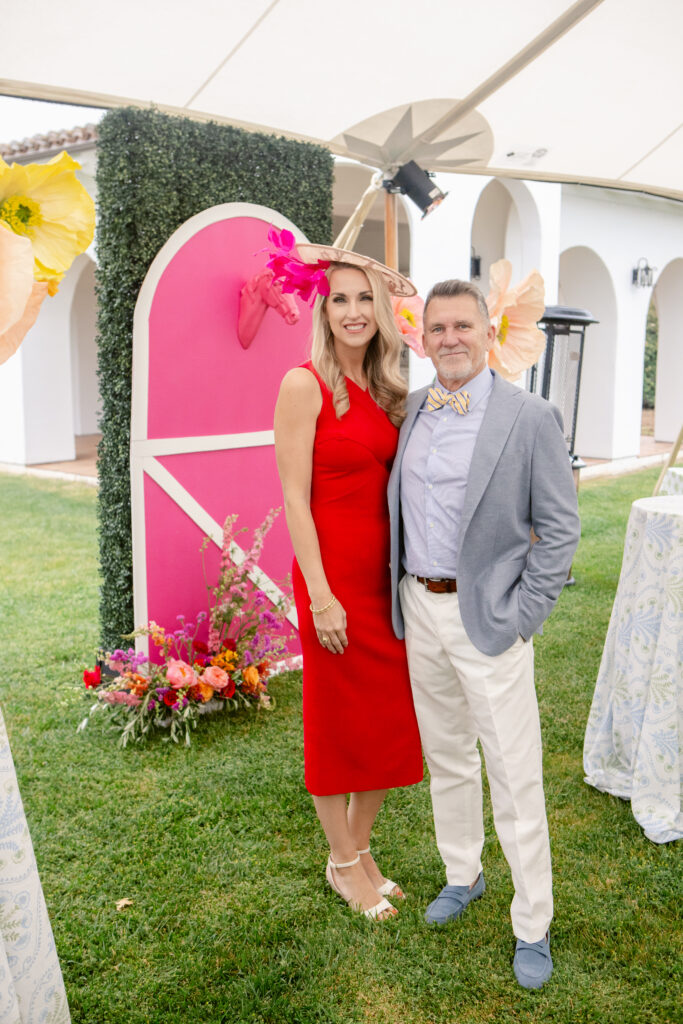 Photo of couple dressed up for the Kentucky Derby party. Photo backdrop of a pink stable door with horse head popping out in background along with tall paper flowers.