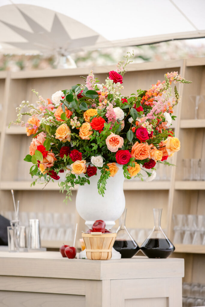 Large floral arrangement with red, orange, and pink flowers in white urn on light wood bar. Accented by a basket of red apples and wine decanters. Large back-bar shelves in background.