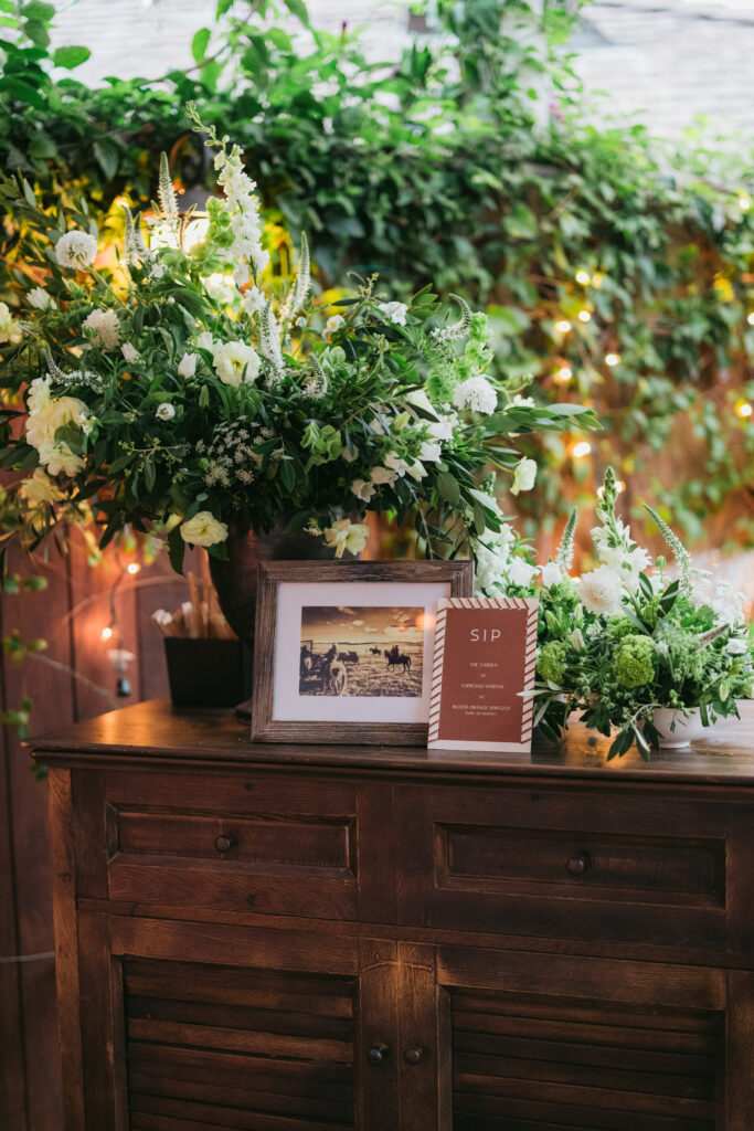 Outdoor wood bar with cozy western vibe. Picture frame with photo of family ranch, large urn floral arrangement with lots of textured greenery and a leather bar sign