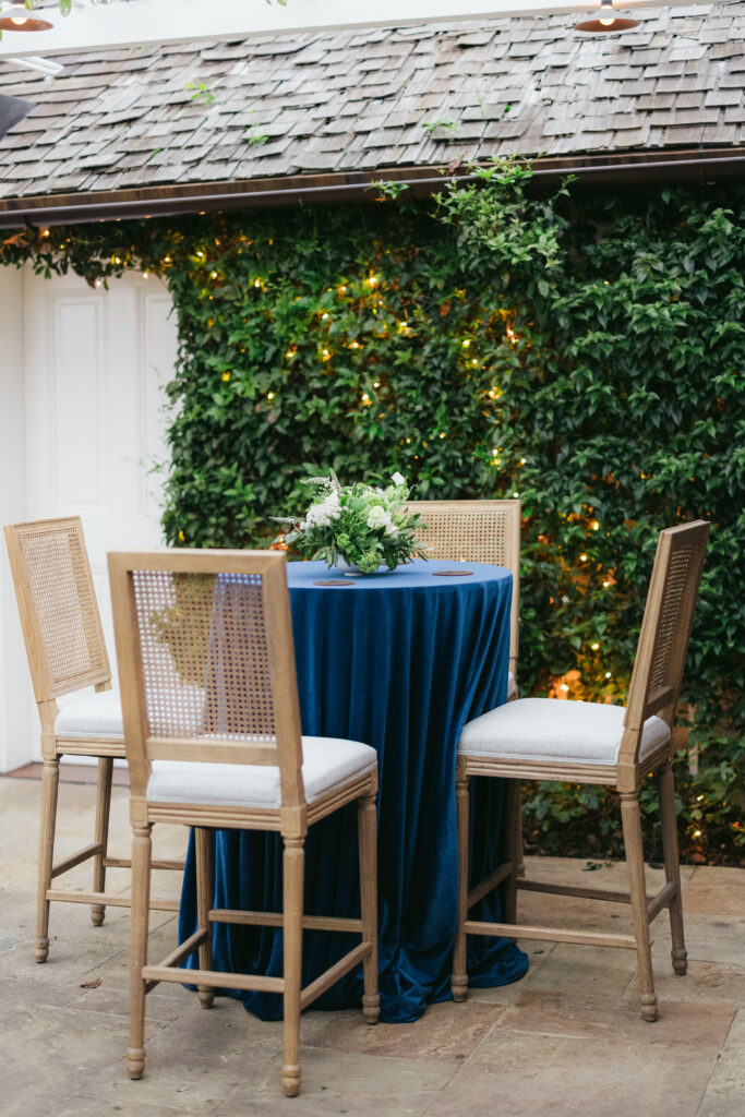 Cocktail table with navy blue velvet linens, green floral centerpiece, and upholstered bar chairs. Outdoor cocktail hour at San Ysidro Ranch